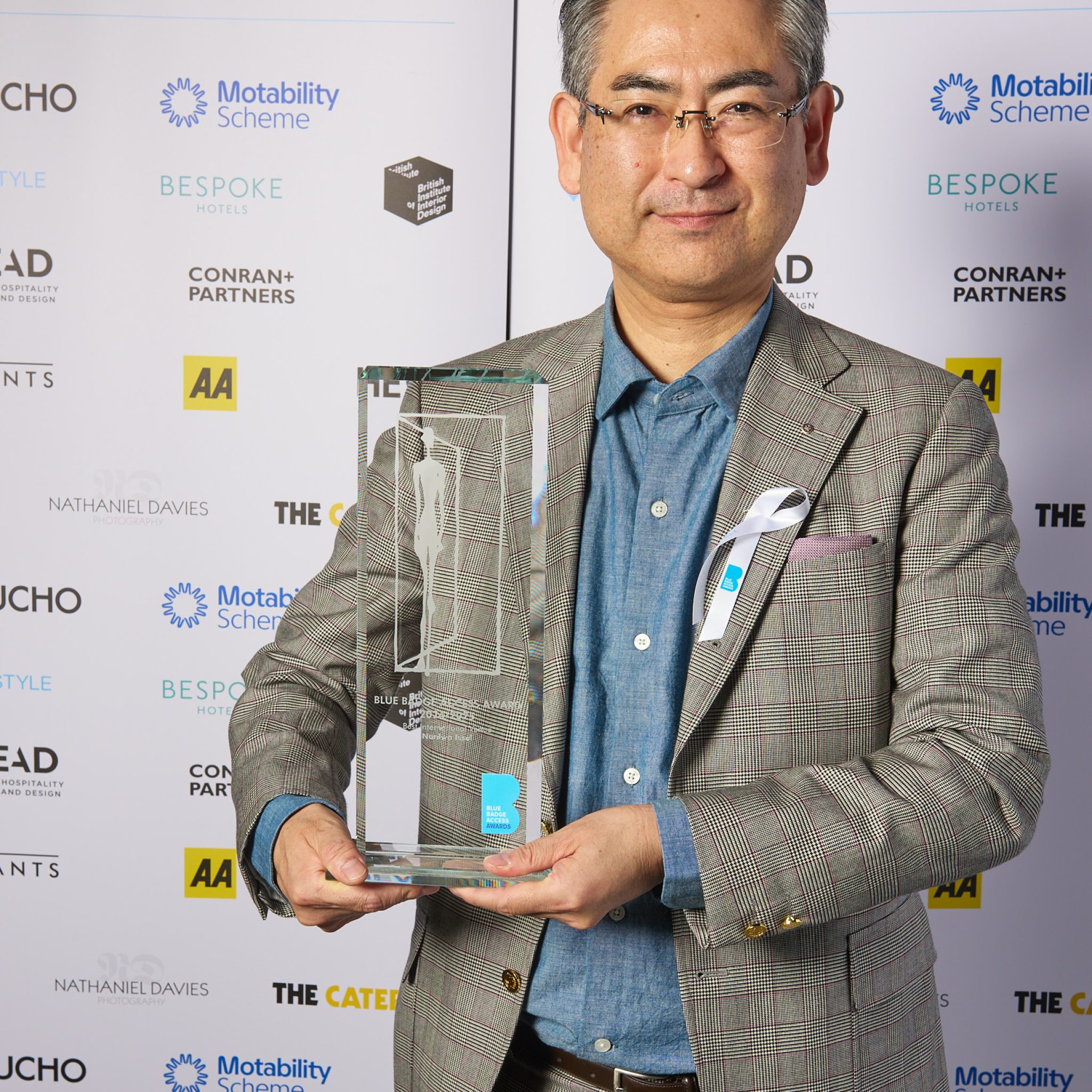 Winner Naniwa Issui holds a Blue Badge Access Awards trophy in front of a sponsor backdrop.