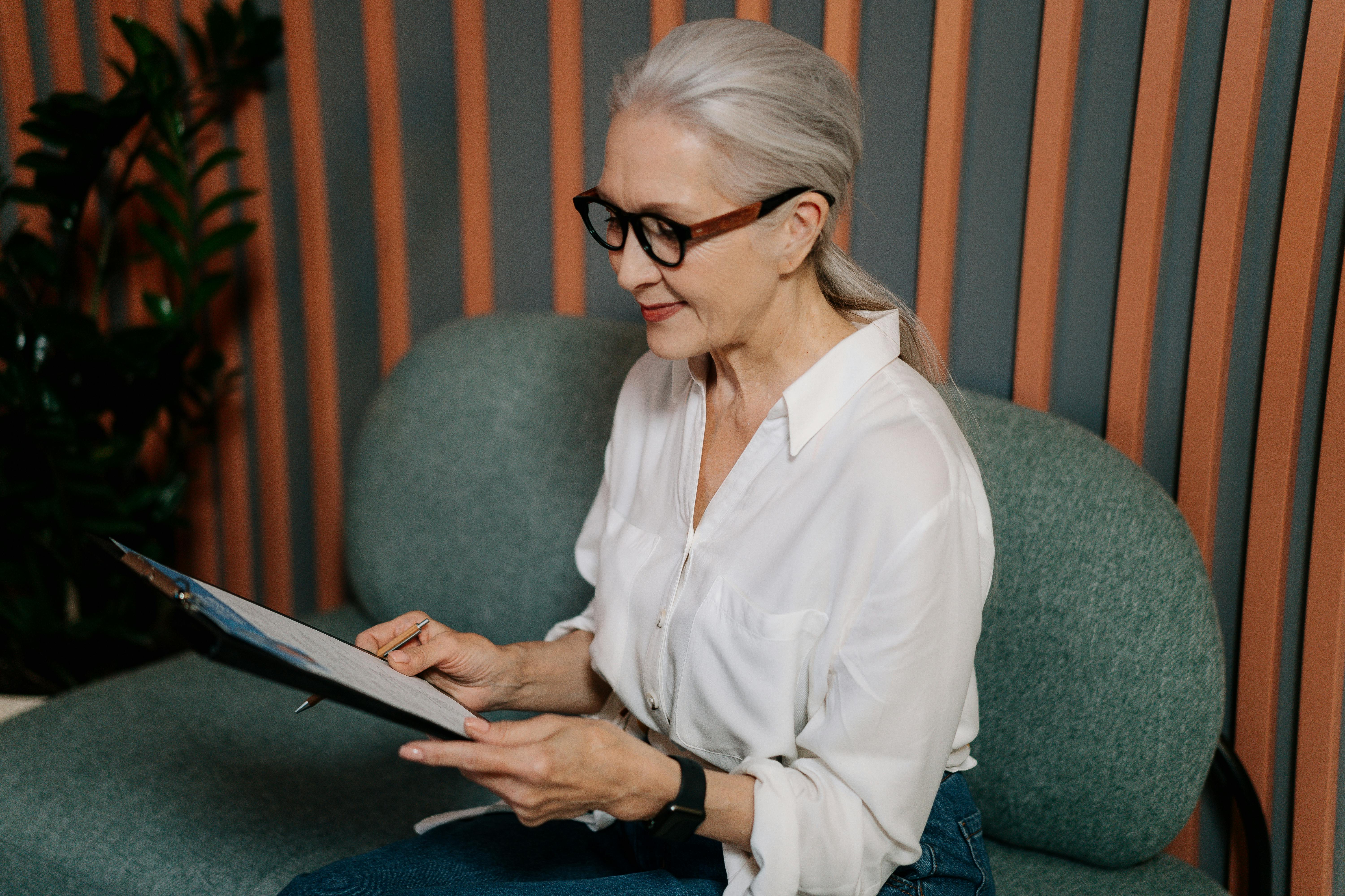 Older woman with grey hair tied back, wearing glasses and a white shirt, sitting on a green chair and reviewing documents on a clipboard, with a striped wall and plant in the background.