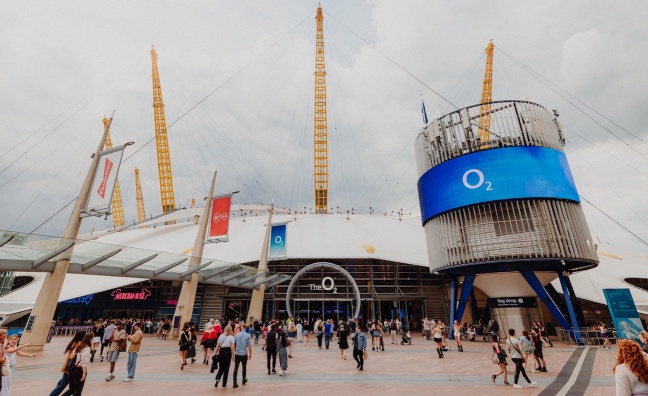 A wide view of the entrance to The O2 arena in London, showing a crowd of people walking towards the venue. The iconic white dome with yellow support masts rises in the background. Large digital screens display the O2 logo, and the surrounding area includes shops and banners. The sky is overcast.