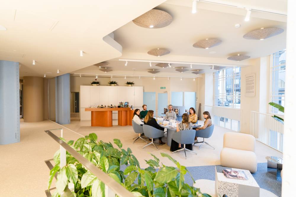 A group of people seated around a circular table in a bright, modern office space, engaged in discussion. The room features soft lighting, acoustic ceiling panels, large windows, and indoor plants.