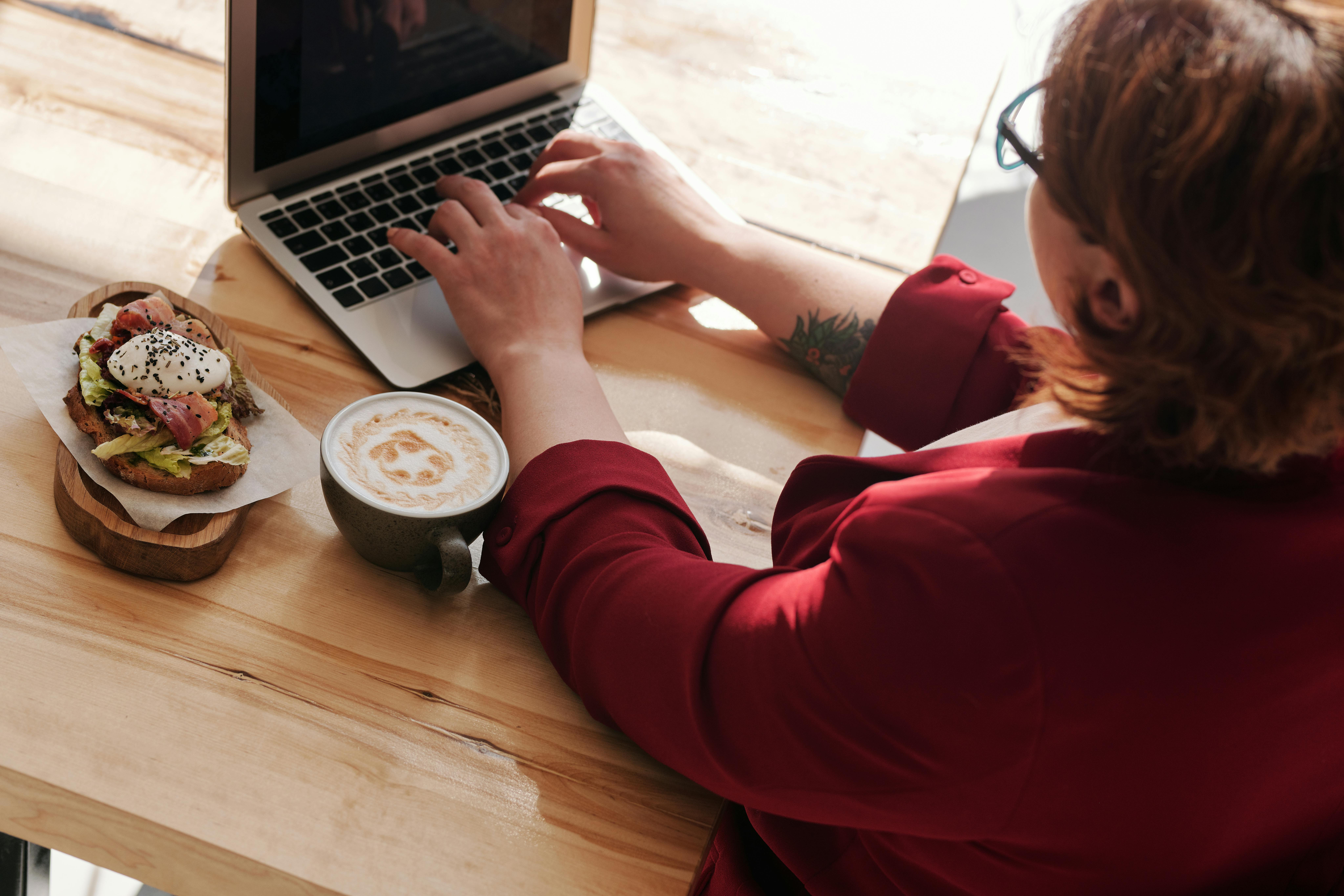 Person wearing a red jacket working on a laptop at a wooden table, with a coffee and an open-faced sandwich topped with lettuce, bacon, and a poached egg nearby.
