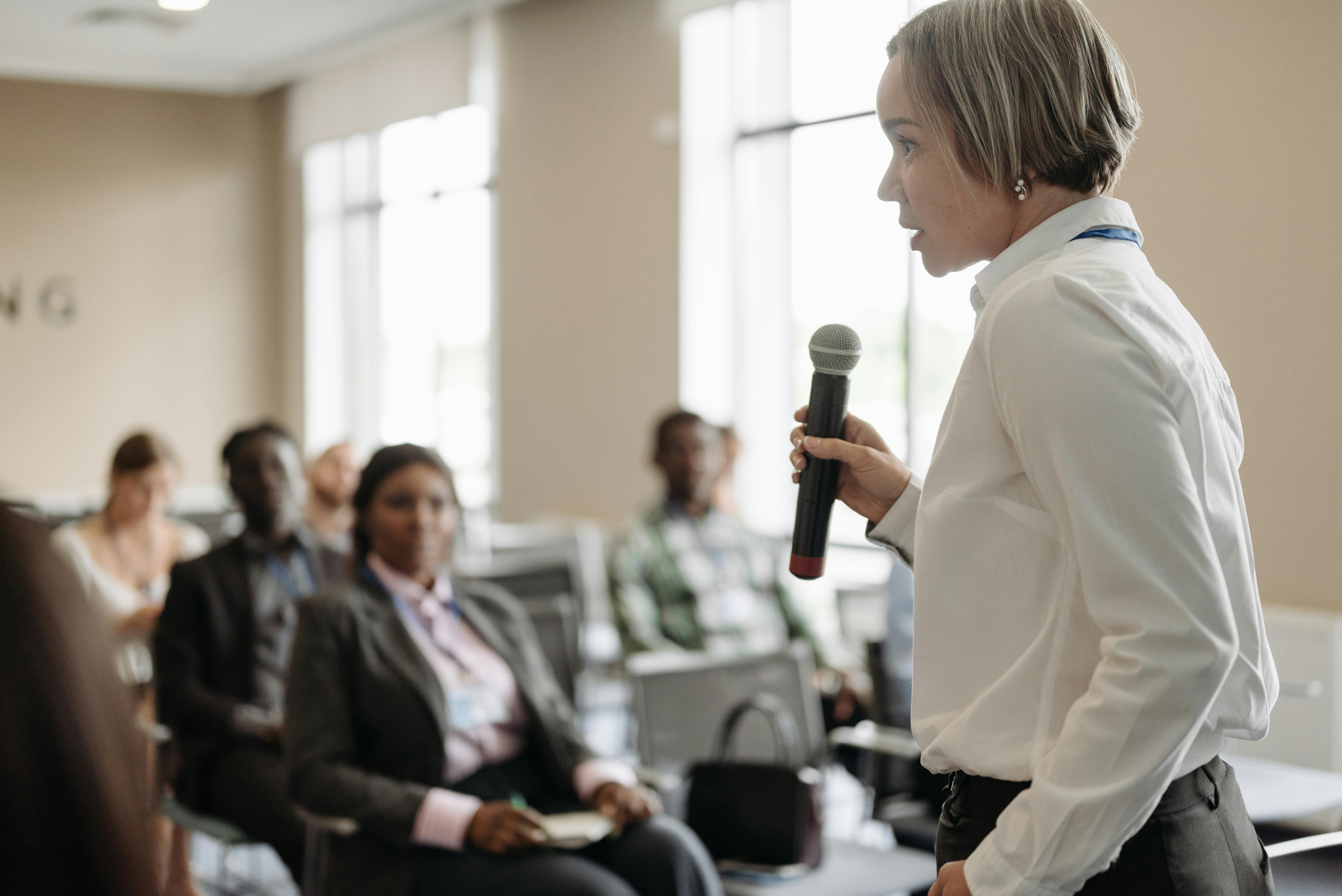 Person holding a microphone and speaking to an audience during a seminar or workshop, with attendees seated and listening in a bright conference room.