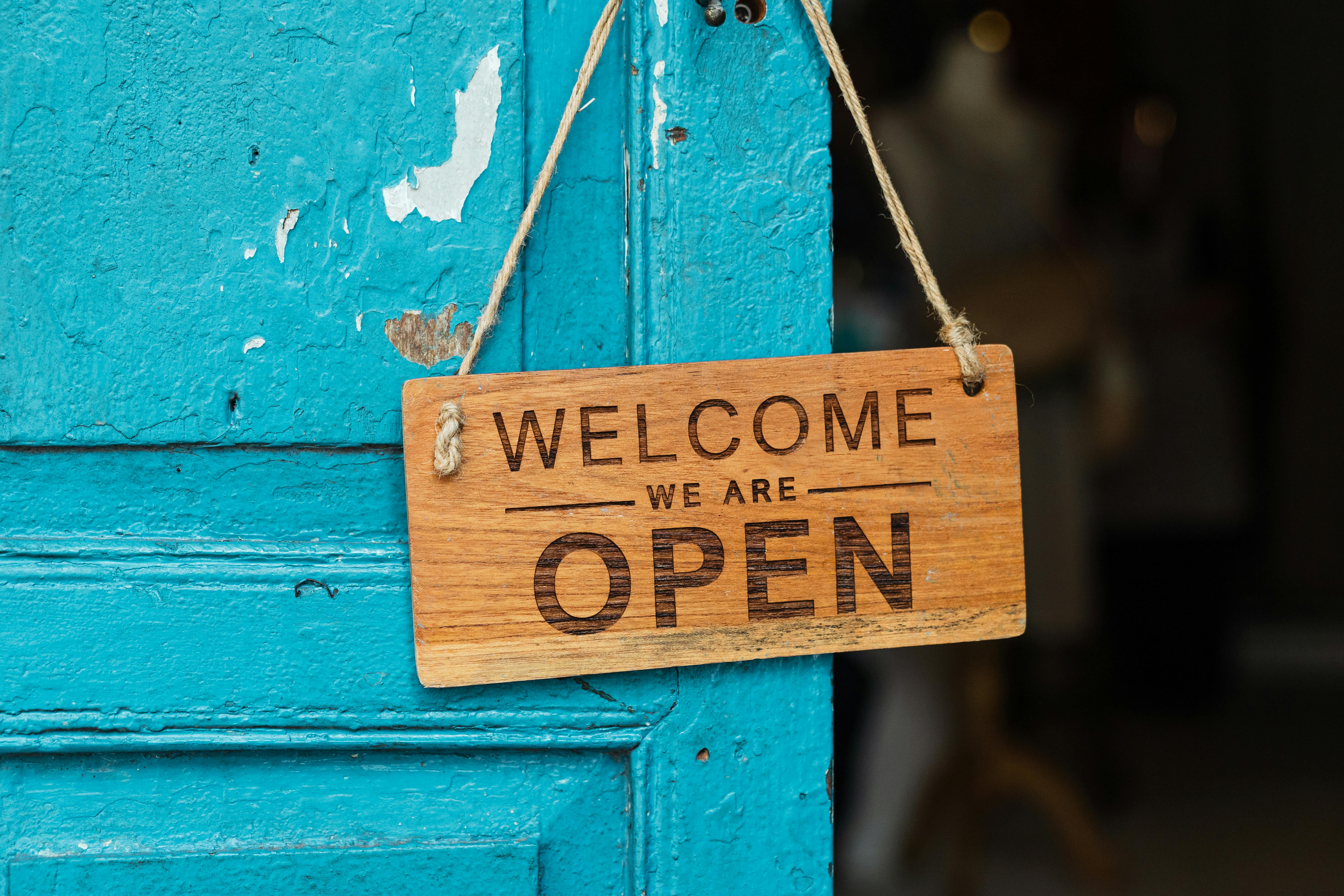 A wooden sign that reads “Welcome, We Are Open” hanging on a bright turquoise door with peeling paint, suggesting a warm and inviting entrance.