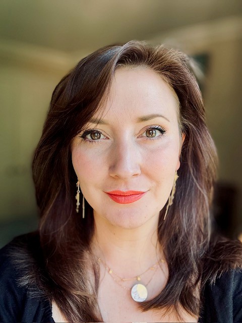 Richmal Maybank, a woman with shoulder-length brown hair and fair skin looks directly at the camera with a slight smile. She is wearing red lipstick, subtle eye makeup, long gold earrings, and two delicate gold necklaces, one with a round pendant. The background is softly blurred, suggesting a room with warm lighting.