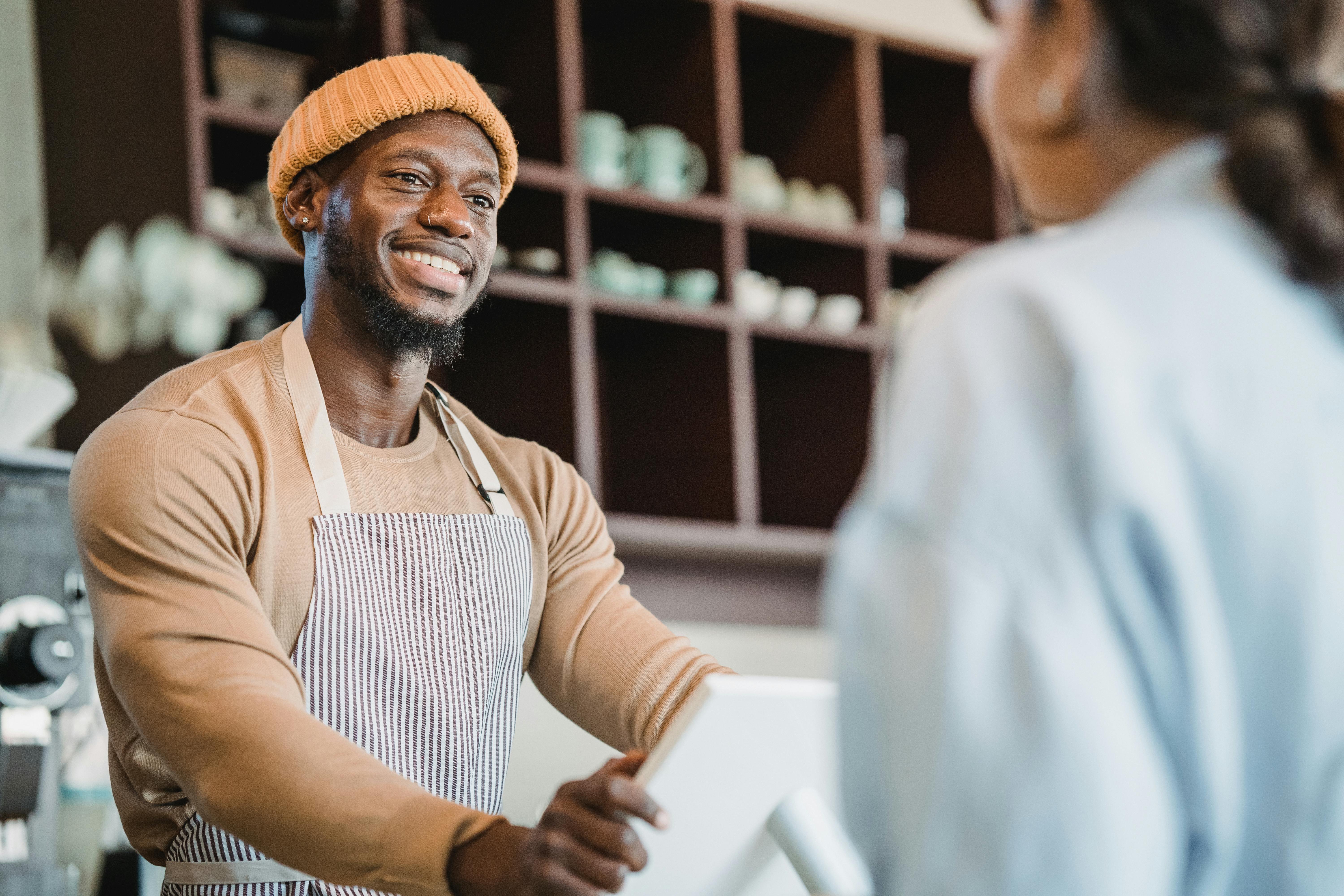 Smiling barista wearing a striped apron and orange beanie serving a customer at a coffee shop counter, with shelves of mugs in the background.