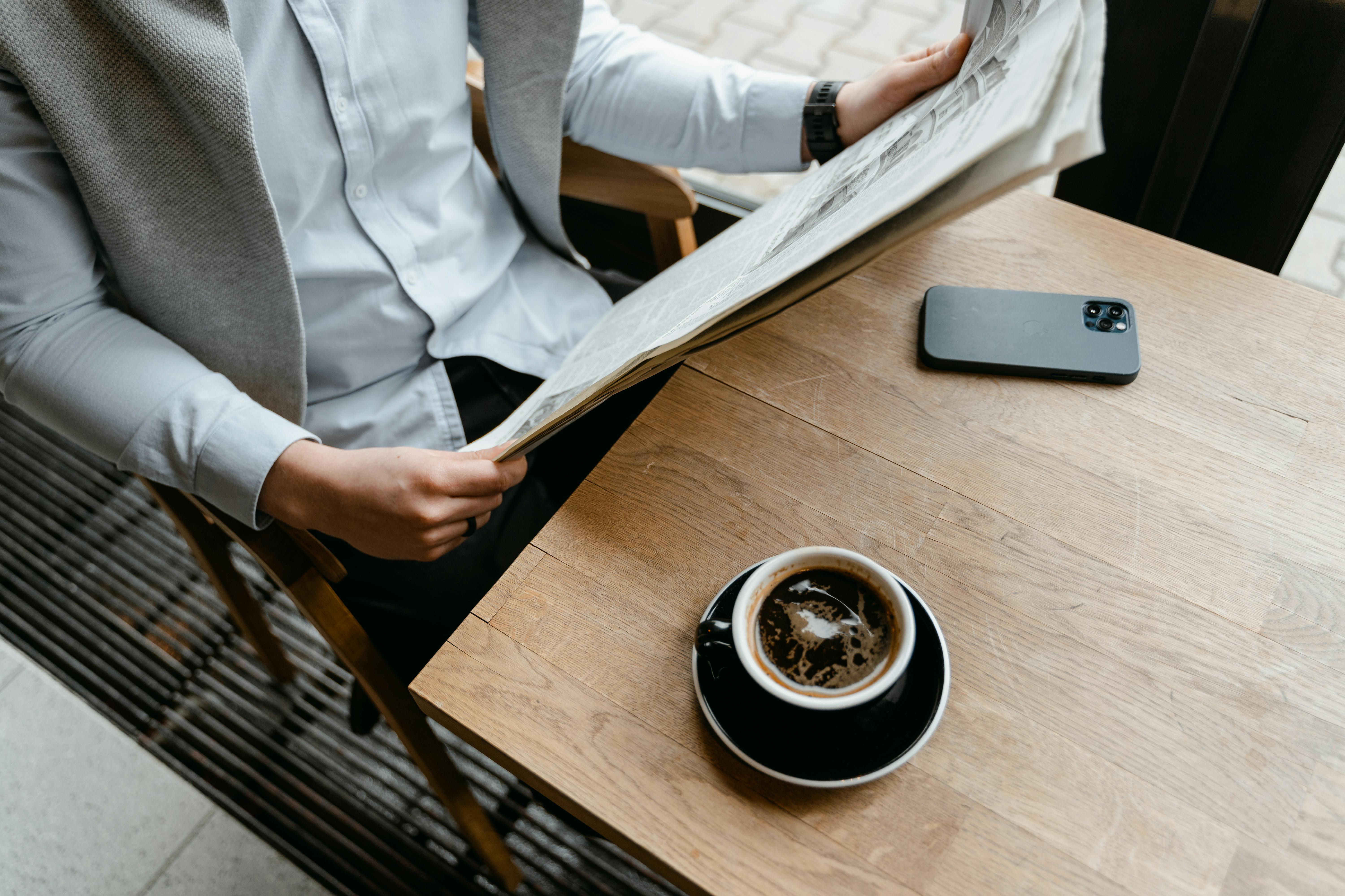 Person sitting at a wooden café table, reading a newspaper with a smartphone beside them and a cup of black coffee in a black saucer in the foreground. The scene suggests a quiet, reflective moment in a relaxed public setting.