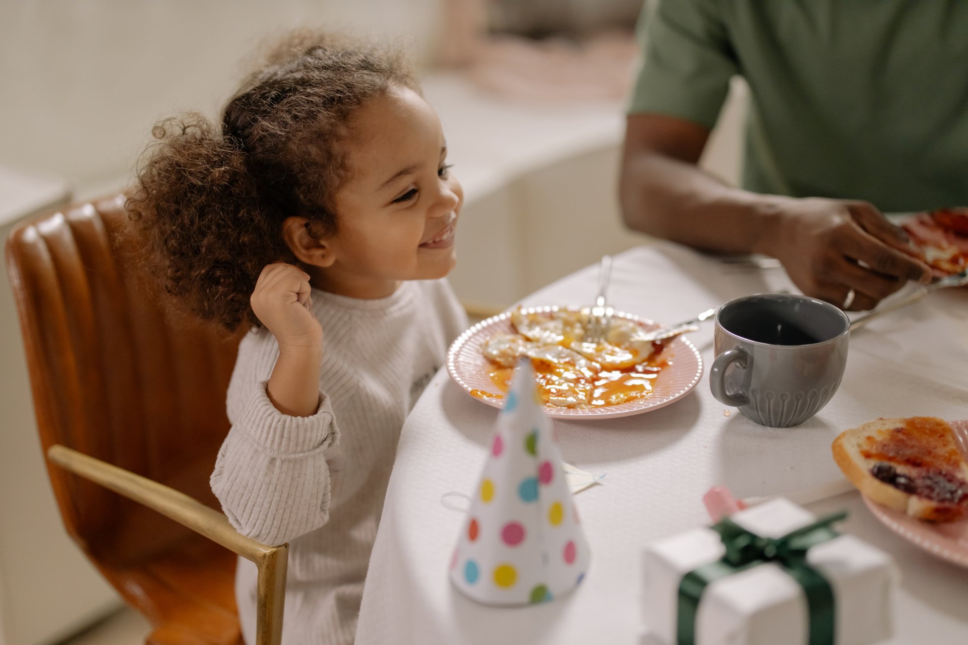 Young child smiling while sitting at a table with a plate of food, a party hat, and a small wrapped gift nearby, sharing a meal with an adult