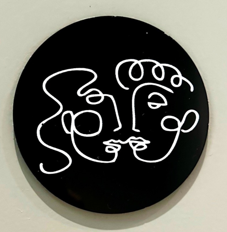 A black circular sign featuring a white abstract line drawing of two faces intertwined, created using a single continuous line.