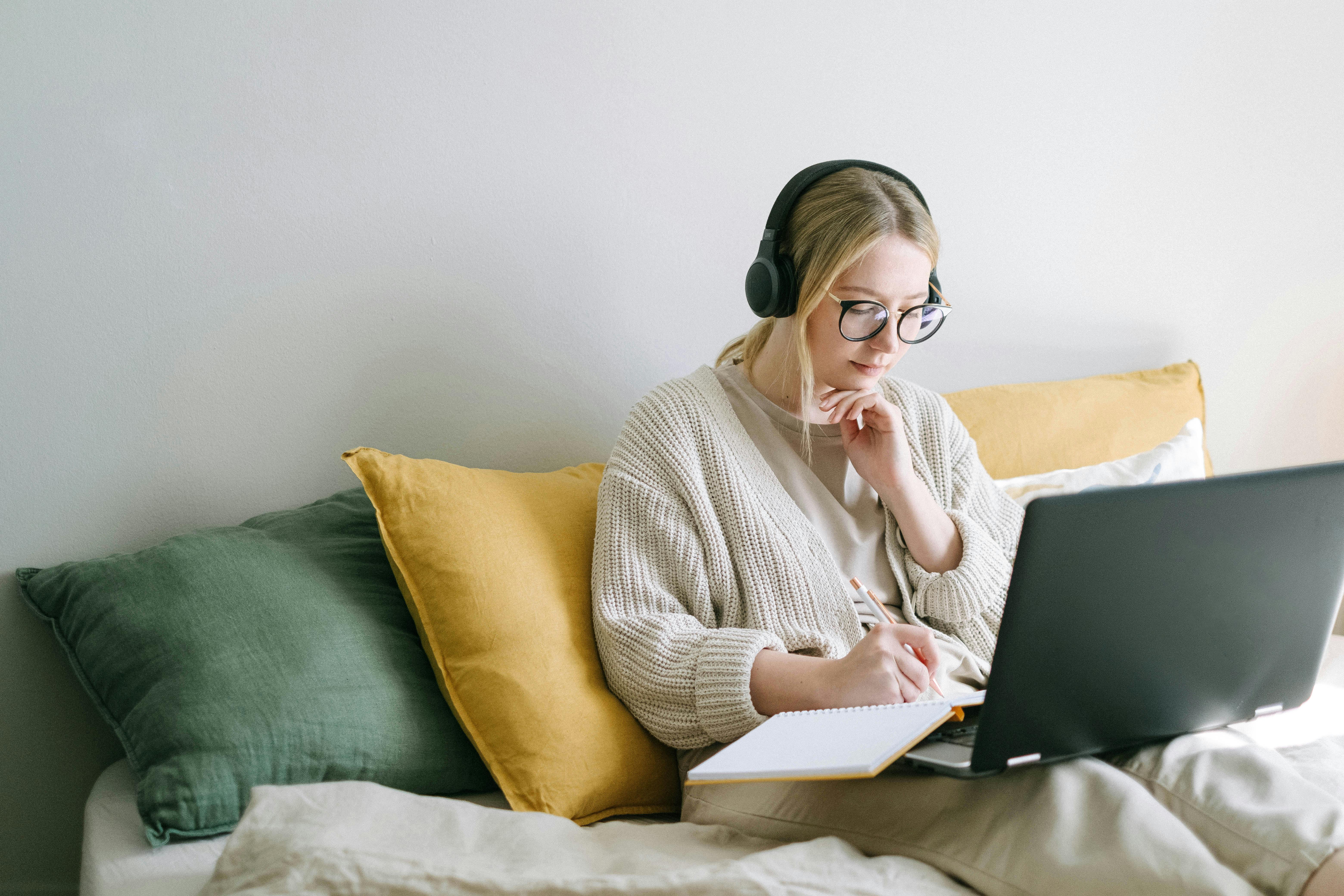 Person wearing glasses and headphones sitting on a bed, writing in a notebook while using a laptop, with green and yellow cushions behind them.