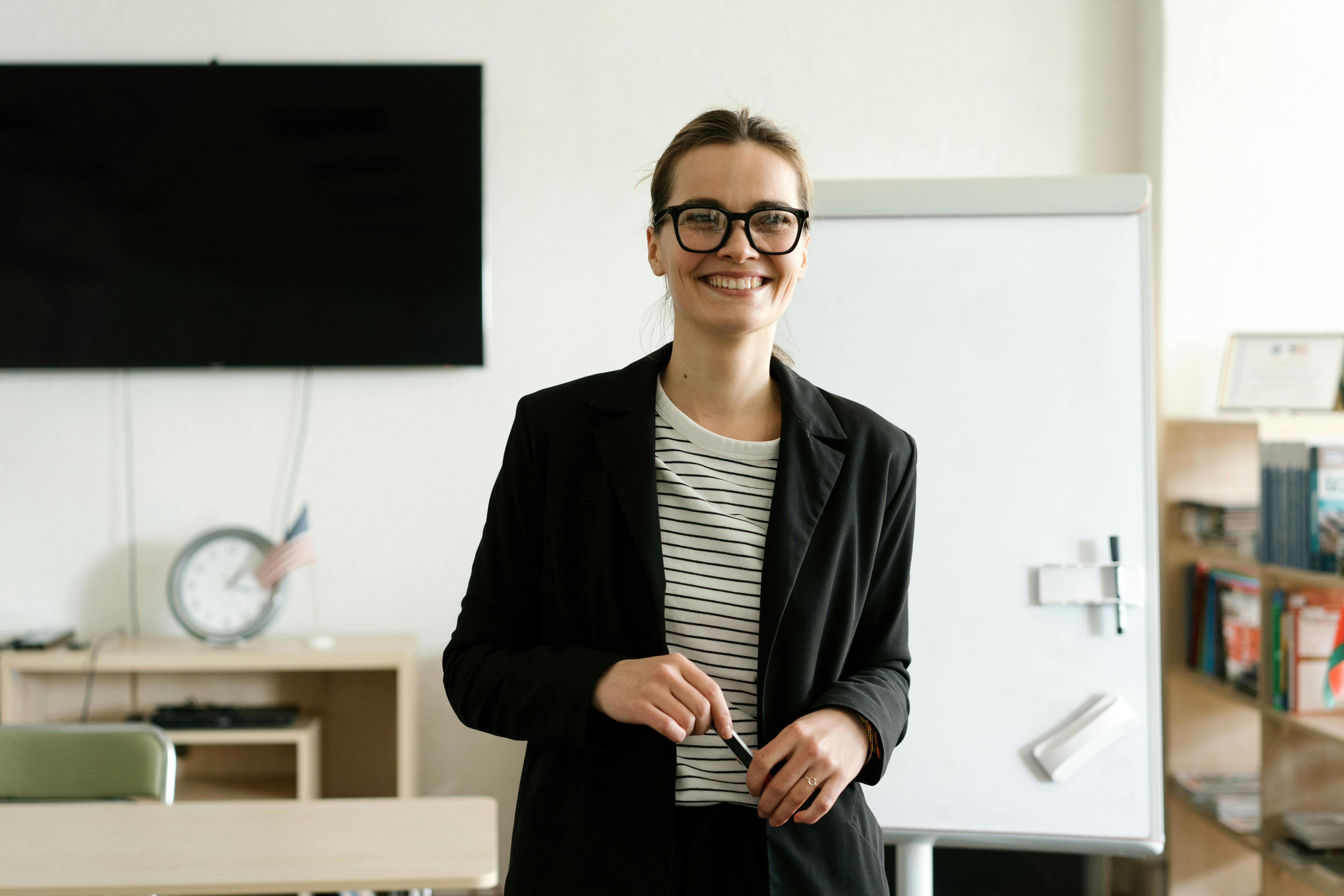 Smiling person wearing glasses, a striped shirt, and a black blazer, standing in front of a whiteboard in a classroom or training room setting.