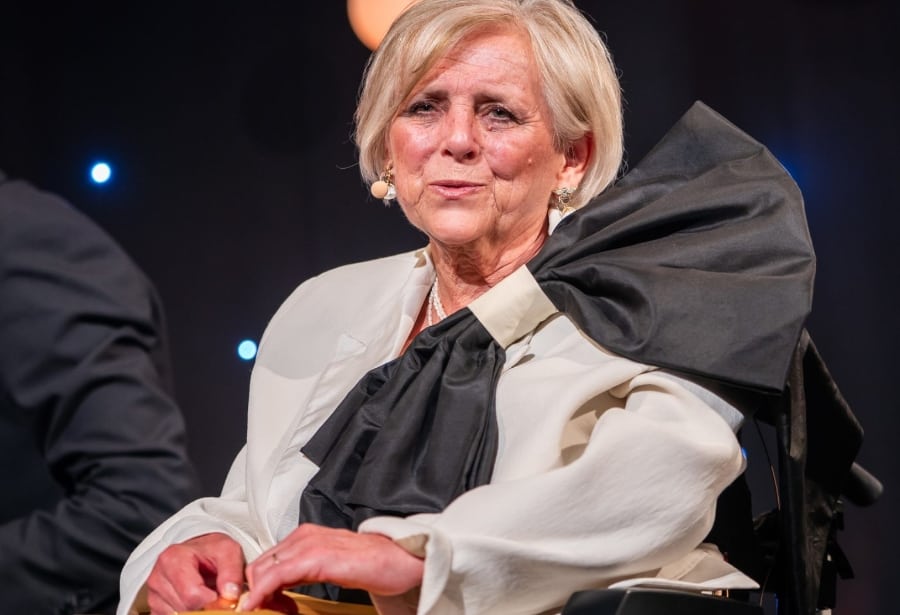 Fiona Jarvis is seated on stage under soft event lighting, wearing a cream blouse with a striking oversized black bow detail at the shoulder. She appears poised and confident, speaking into a discreet headset microphone. The background is dark with subtle stage lights, suggesting a formal awards or conference setting. Fiona is seated in a wheelchair and is positioned at the centre of attention, conveying authority and presence.