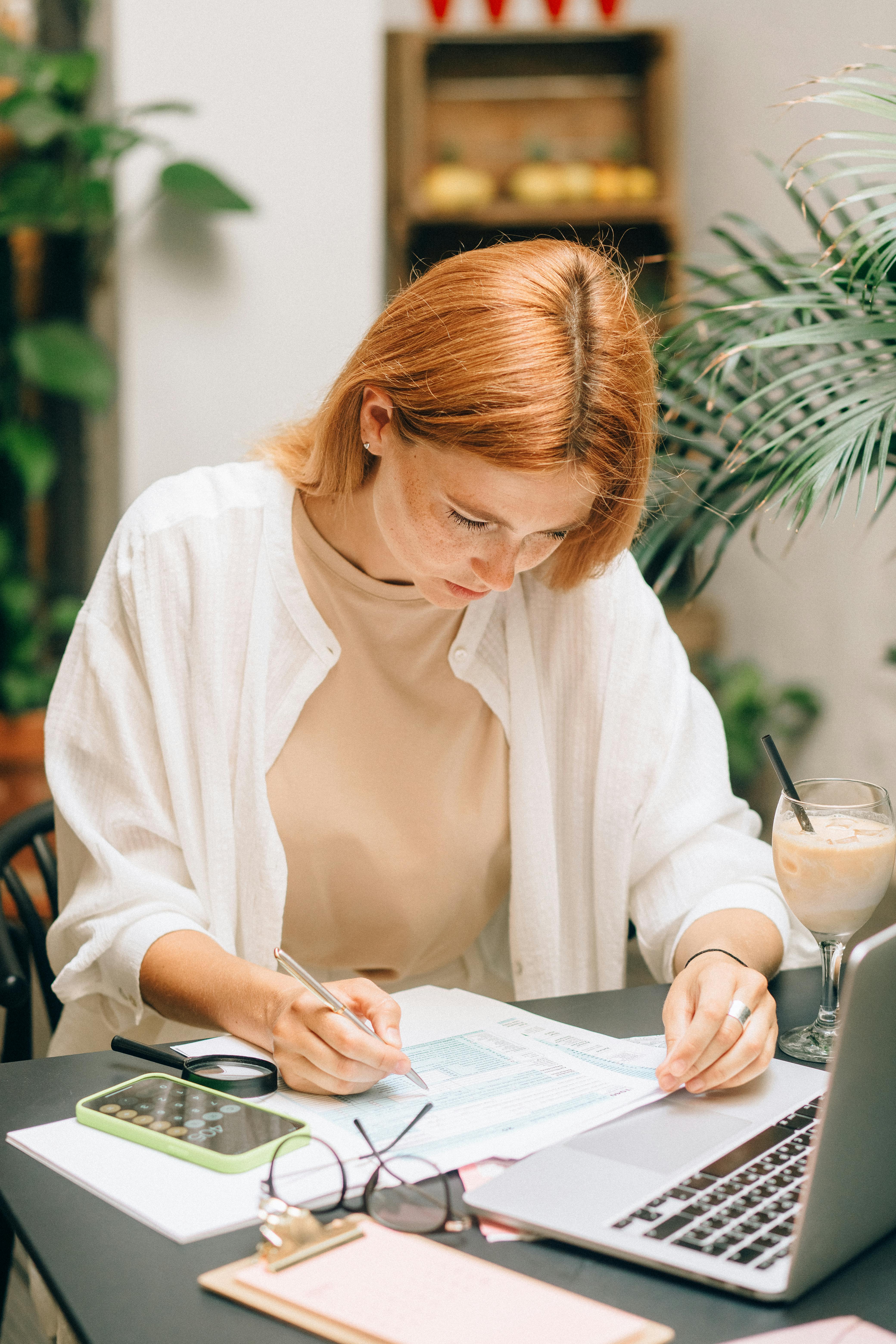 oung woman with red hair sitting at a table, filling out paperwork with a pen next to a laptop, calculator, glasses, and an iced coffee, in a bright space with plants.