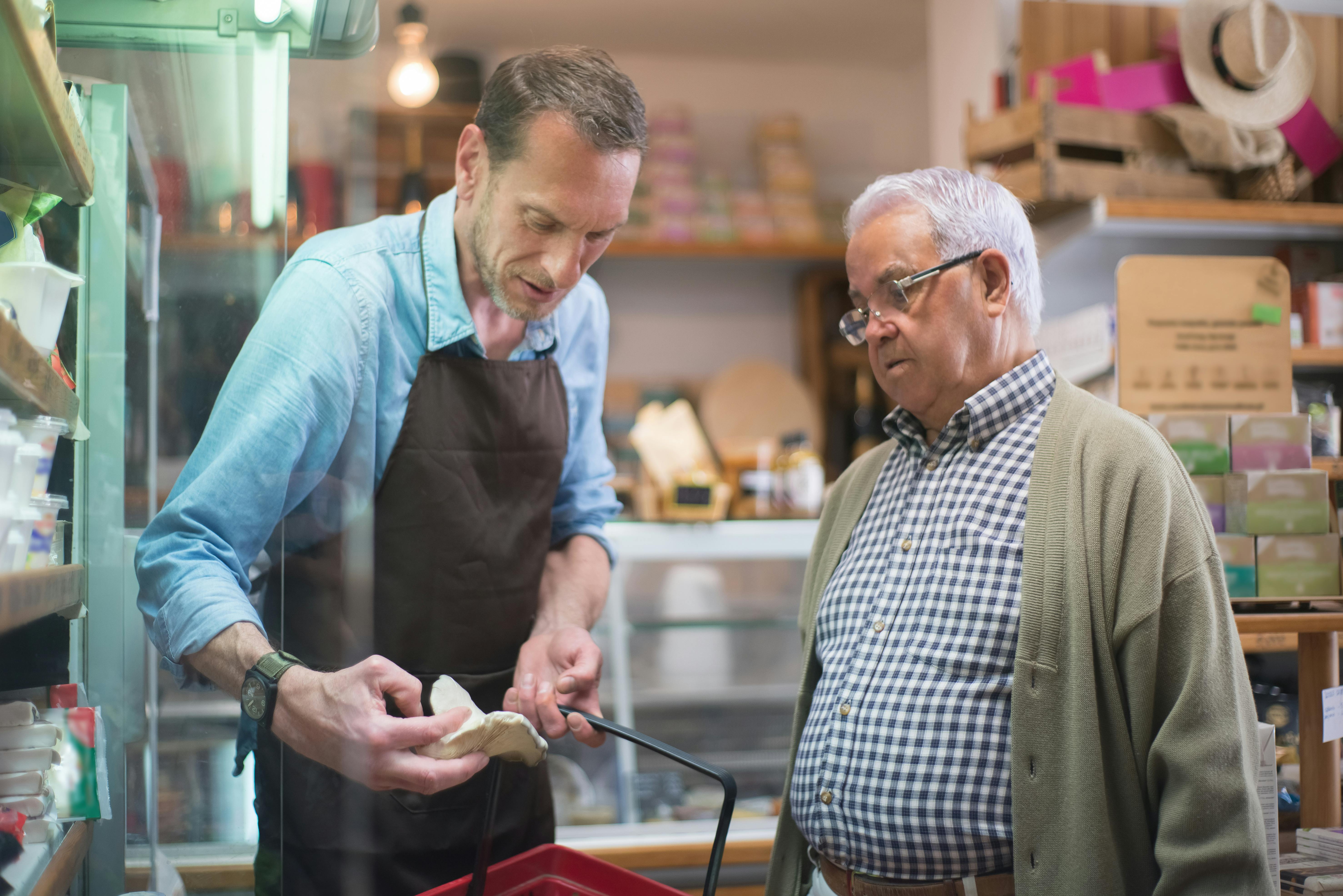 Shop assistant wearing an apron helping an older customer at a counter in a small shop, with shelves and products in the background.