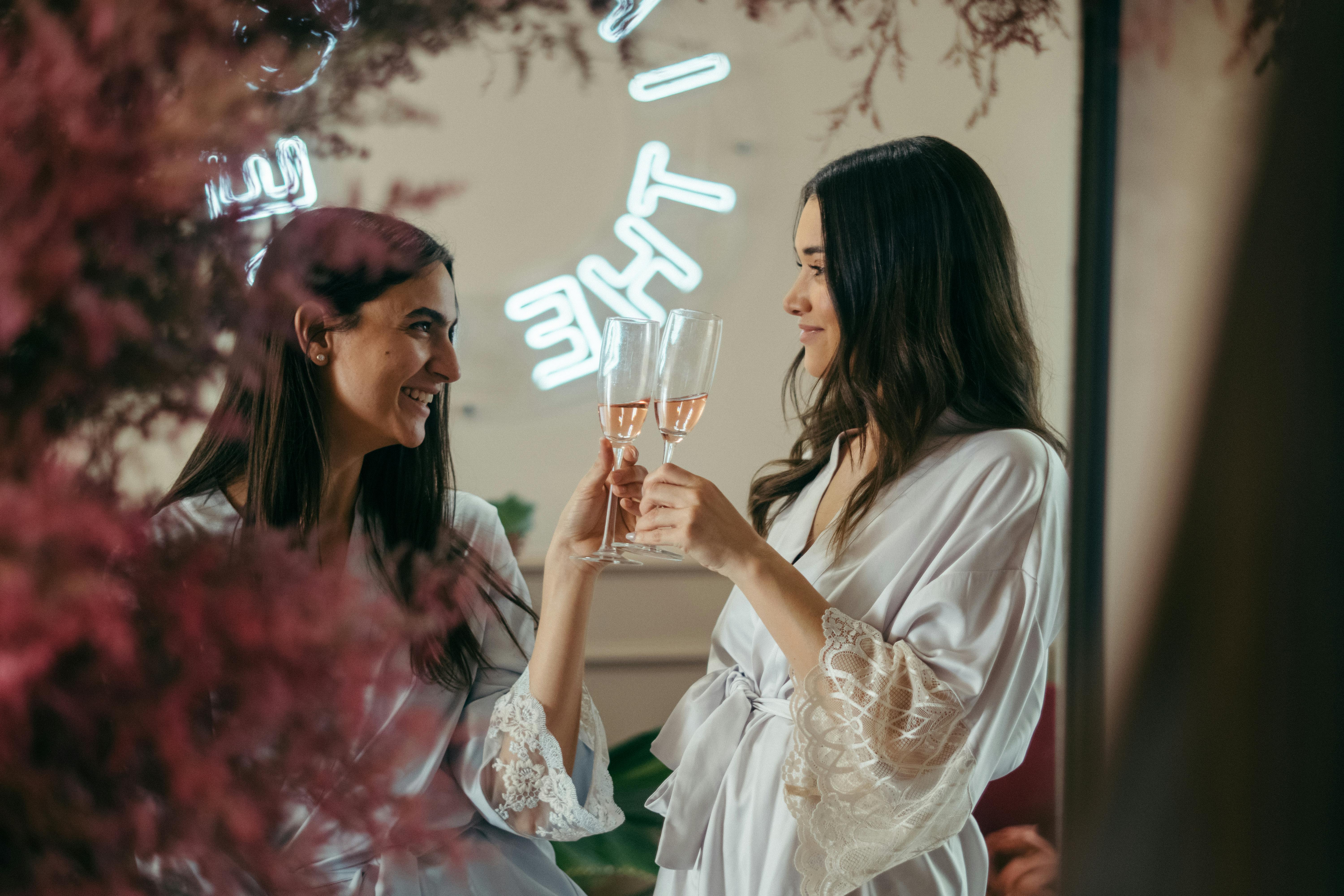 Two women wearing light-coloured robes smile at each other while clinking glasses of rosé champagne. They are standing in front of a mirror with soft lighting and pink foliage in the foreground. A neon sign is partially visible in the background.