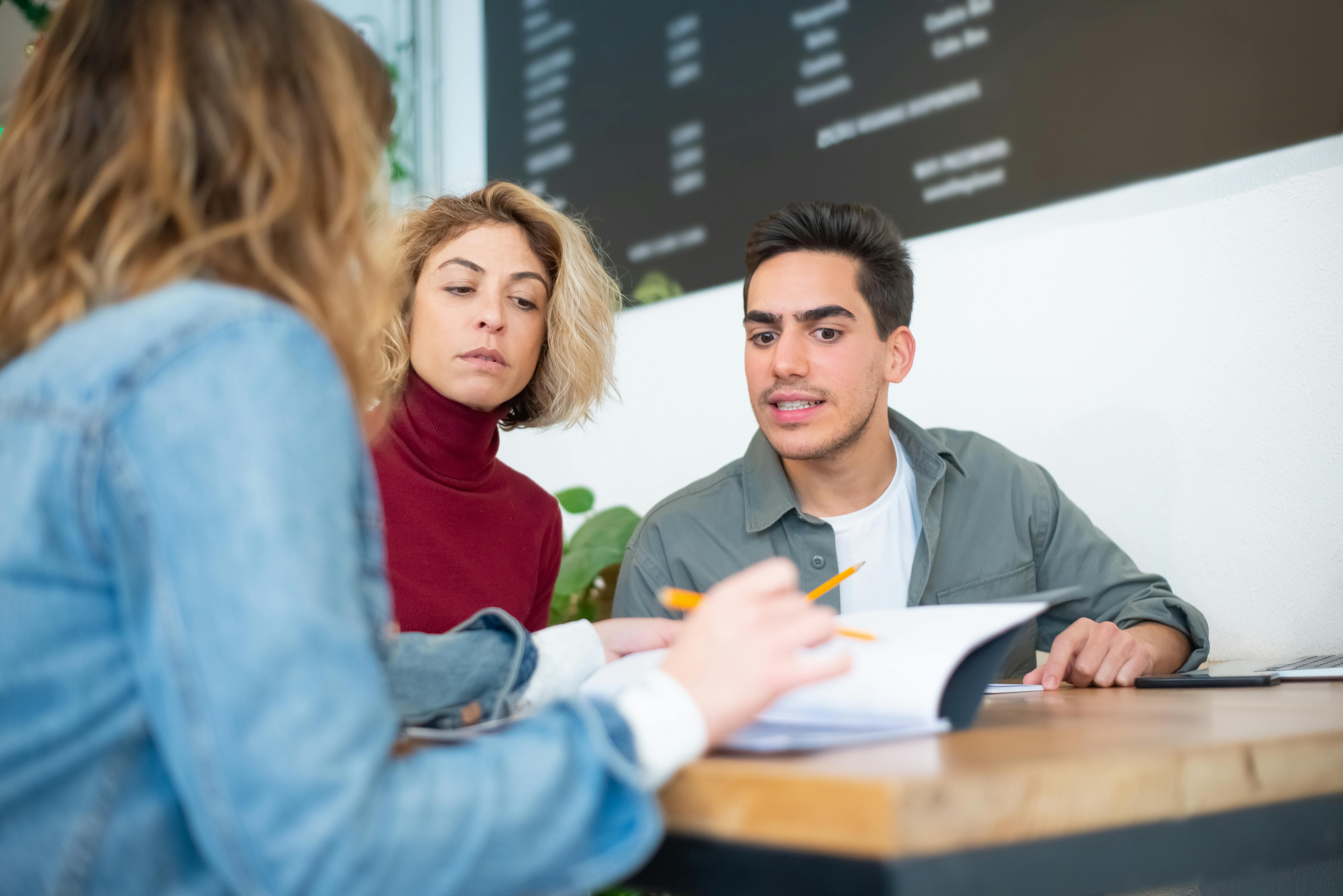 Group of three people sitting at a table, discussing and reviewing documents together, with one person pointing at paperwork while the others listen attentively