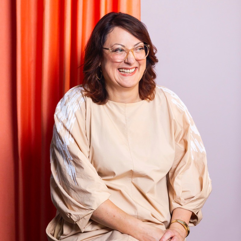 A picture of Justine Fox, wearing glasses and a white dress with background of a pink wall and bright orange curtain