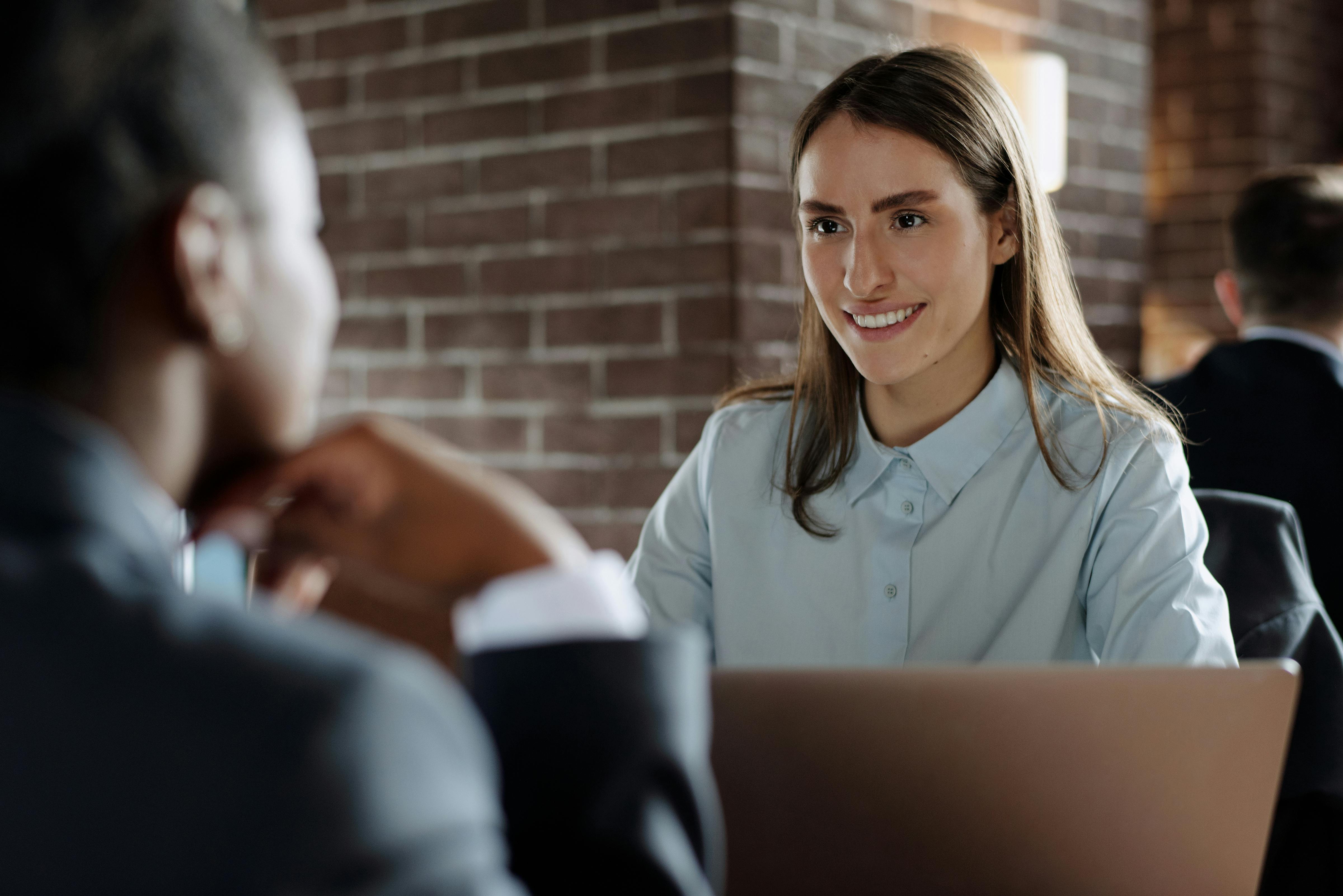 Two people having a conversation across a table in a professional setting, with one smiling woman facing the camera and a laptop in front of her, against a background of exposed brick walls