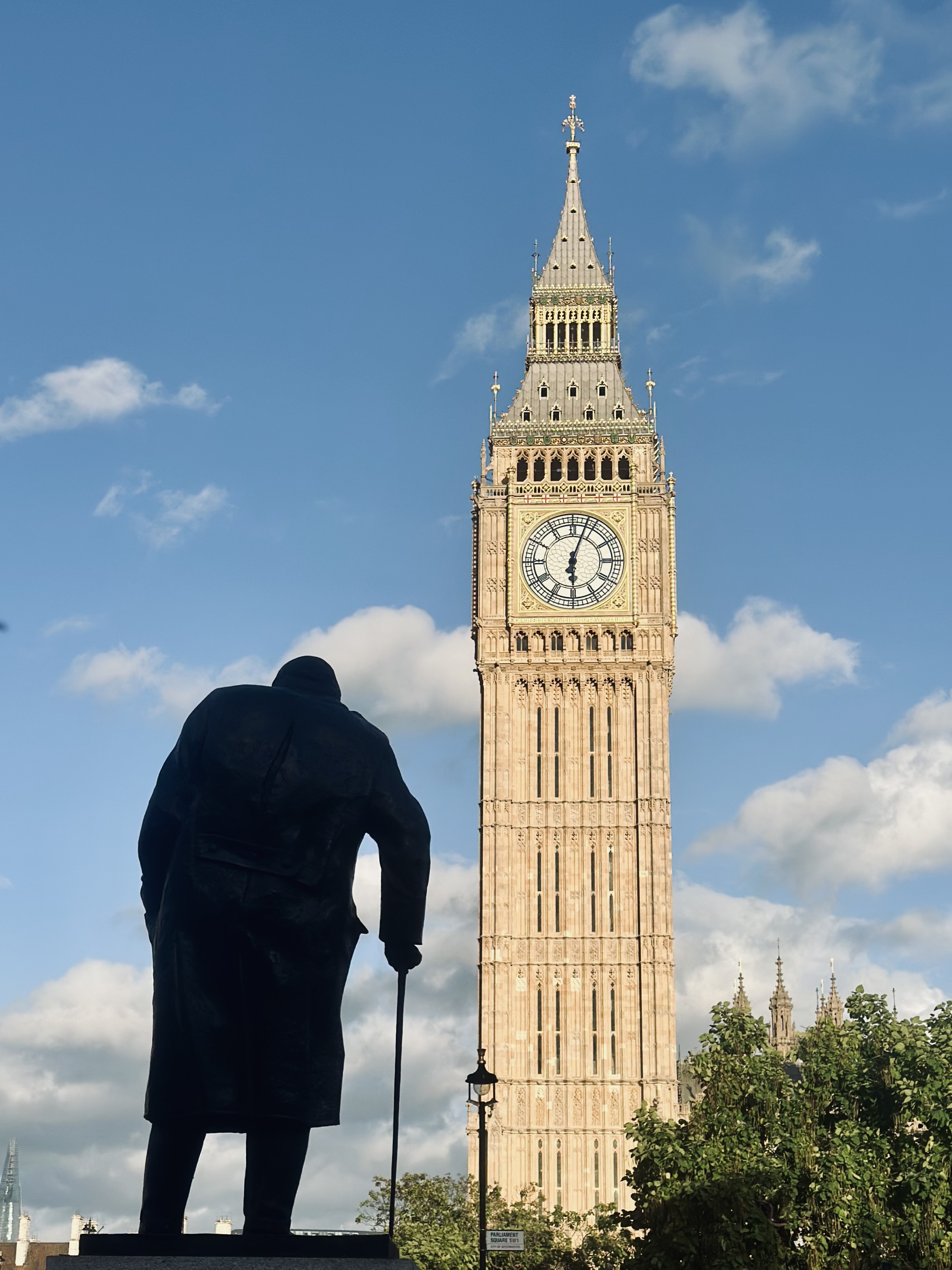 A bronze statue of Winston Churchill stands in silhouette with his cane in Parliament Square, looking towards the Elizabeth Tower (Big Ben) against a bright blue sky with scattered clouds.